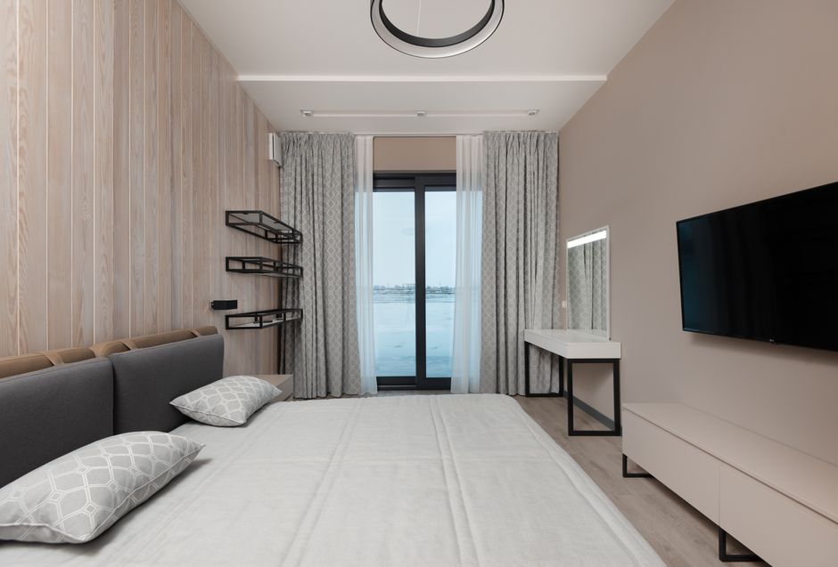 Soft bed with cushions and modern TV hanging on wall over cabinet in minimalist bedroom with vanity table and curtains