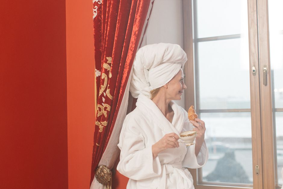 Woman in bathrobe with coffee and croissant looks out window, enjoying a leisurely morning