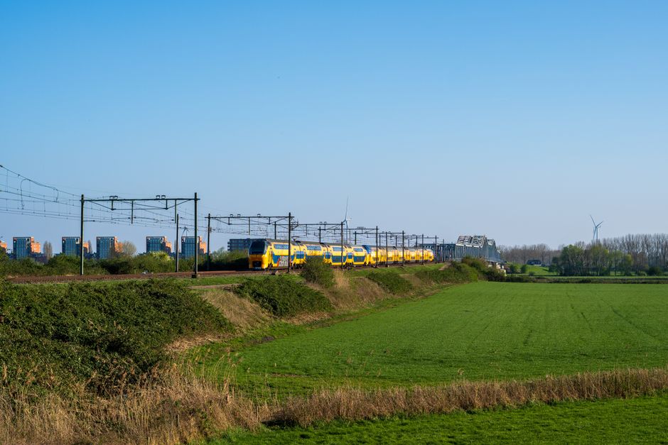 Yellow train crossing a rural area with green fields and distant city buildings under a clear blue sky