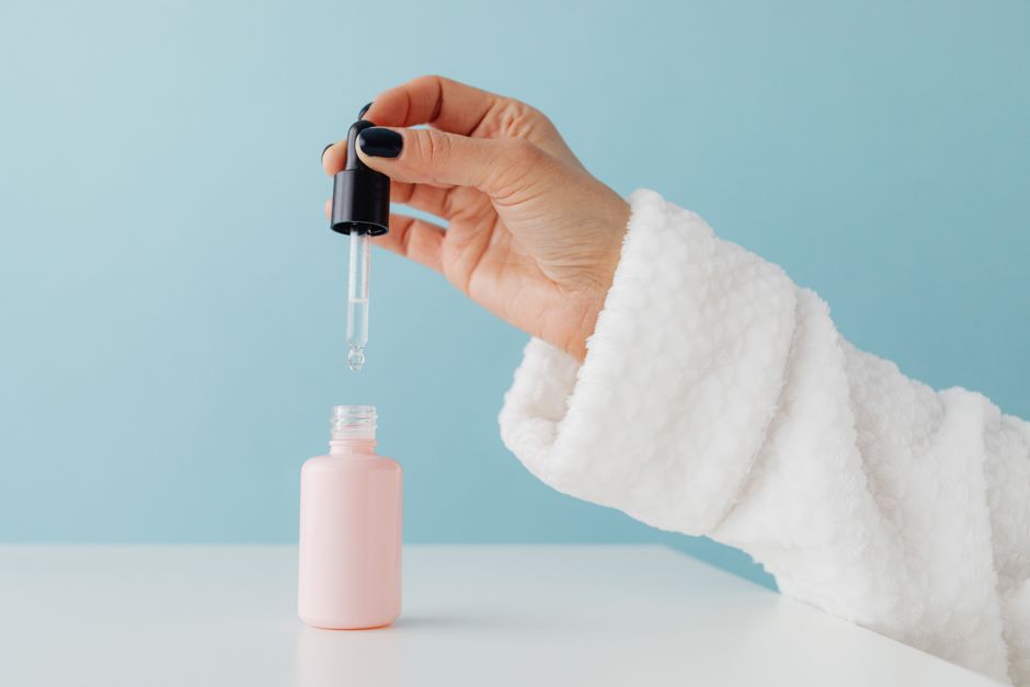 Close-up of a woman's hand in a bathrobe using a serum dropper over a pink glass bottle against a blue