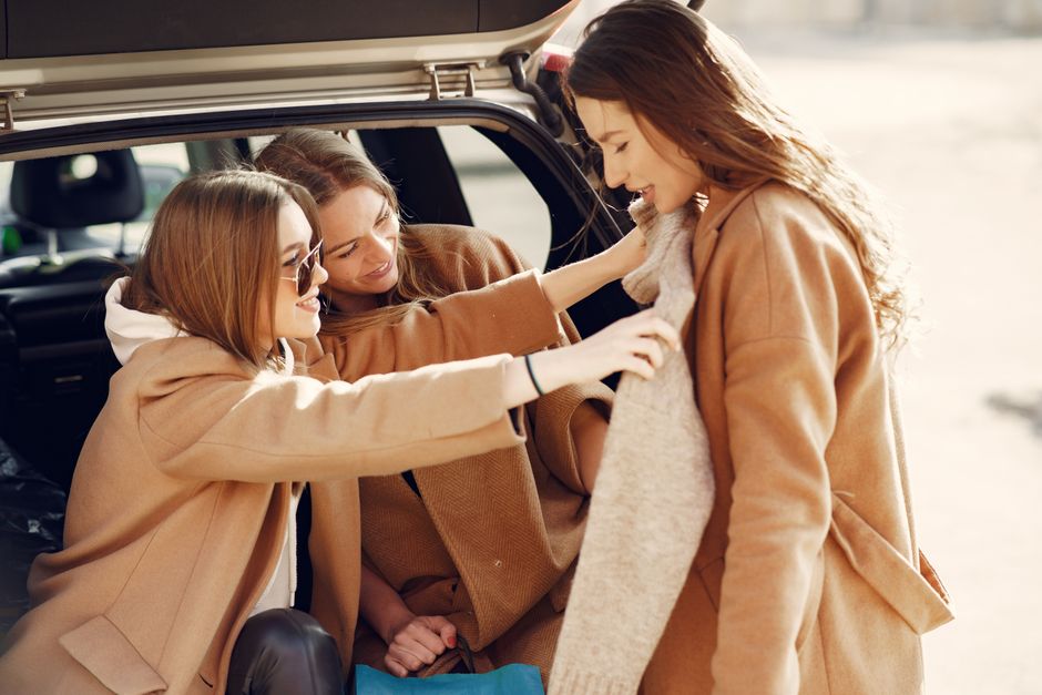 Two young women trying on a friend's new sweater while sitting in a car boot