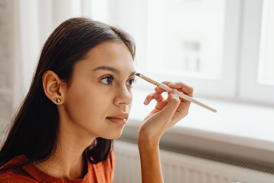 A young woman skillfully applying makeup with a brush by a window indoors