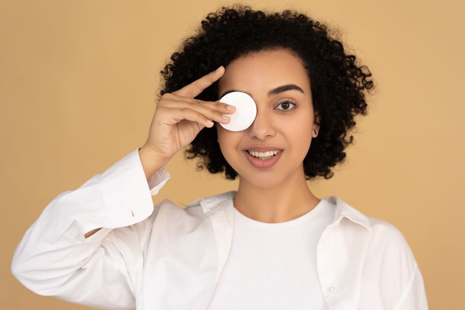 Smiling woman holding a cotton pad against her eye, enjoying skincare routine in a studio setting