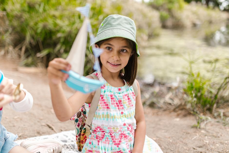 A young girl in a colorful dress and bucket hat playing with a toy boat outdoors