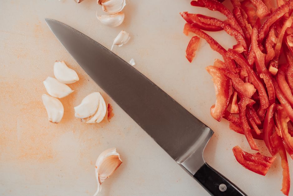 Top view of a kitchen knife with sliced red peppers and garlic on a chopping board