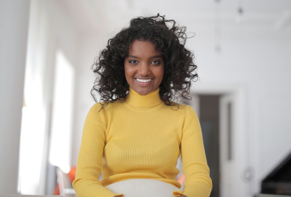 Cheerful young ethnic lady in casual yellow sweater sitting on comfortable seat in light living room and smiling while looking