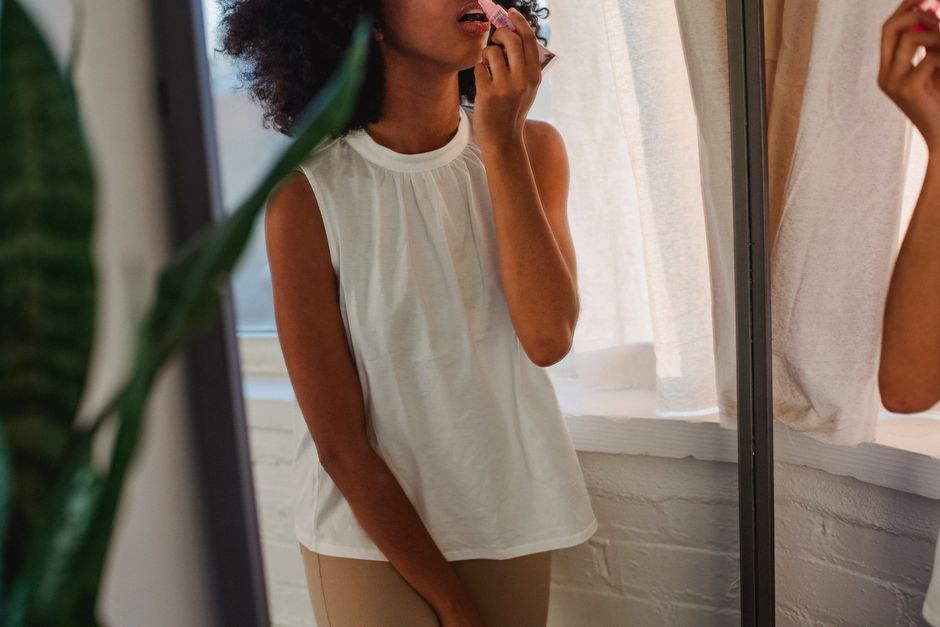 Young African American female in casual clothes standing against big mirror and applying lip gloss at home